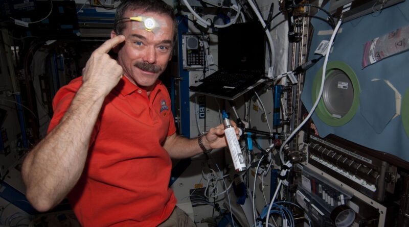 Hadfield is wearing an orange polo shirt and khaki pants and holding a silver canister with a white label and blue tip in his left hand. With his right hand, he is pointing to a round yellow sensor taped to his forehead. There is a laptop over his left shoulder and multiple cords, wires, and switches on the wall in front of him.